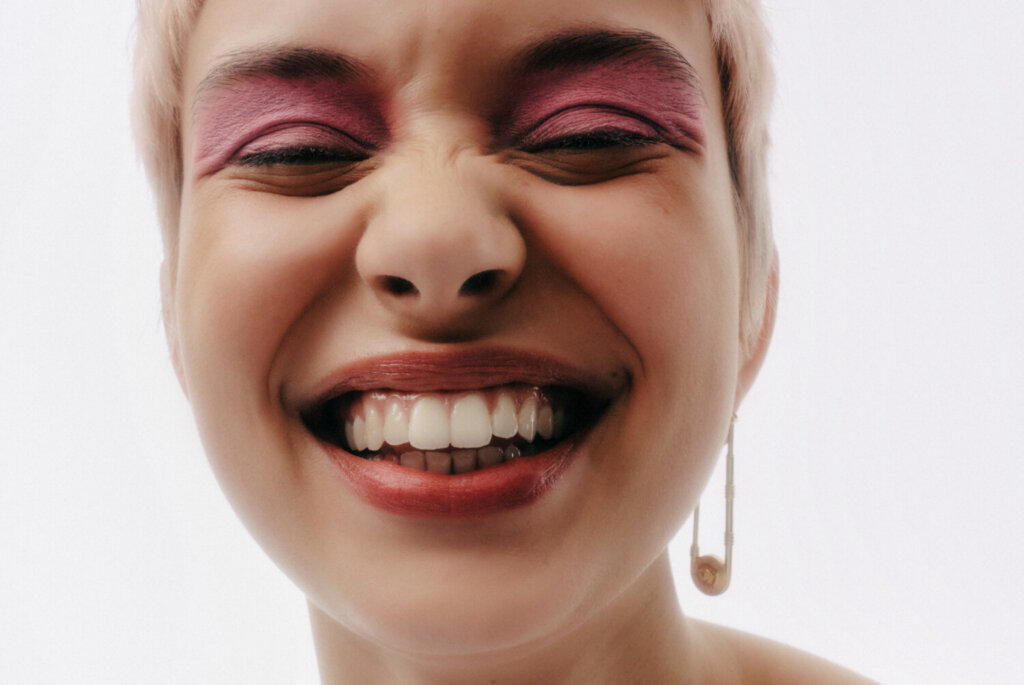 Close-up portrait of a person with eyes closed, bold pink eyeshadow, and a wide, open smile.